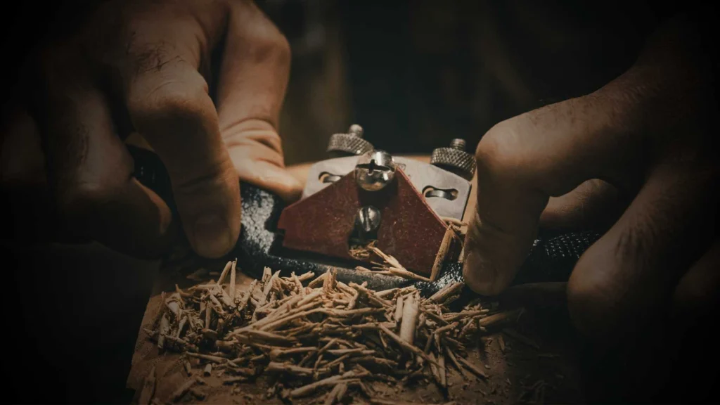 Boutique woodworking craftsman hand planing timber with a spokeshave tool inside a dark custom OEM ODM wood wall panels manufacturing facility.